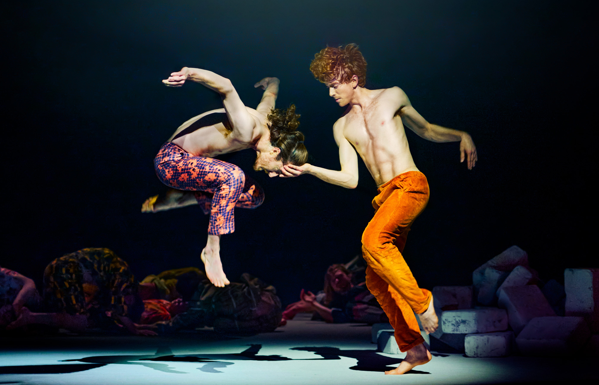 Two individuals in a dynamic and expressive dance pose. They are surrounded by darkness, save for a beam of light that illuminates them and casts shadows on the floor. One person is dressed in orange pants, while the other wears patterned pants. The background is dark, highlighting the dancing figures. Shadows on the floor mirror the dynamic poses of the two individuals. The light is focused on the dancers, providing the image with a dramatic effect. There are multiple objects or people in the background that are not clearly visible.