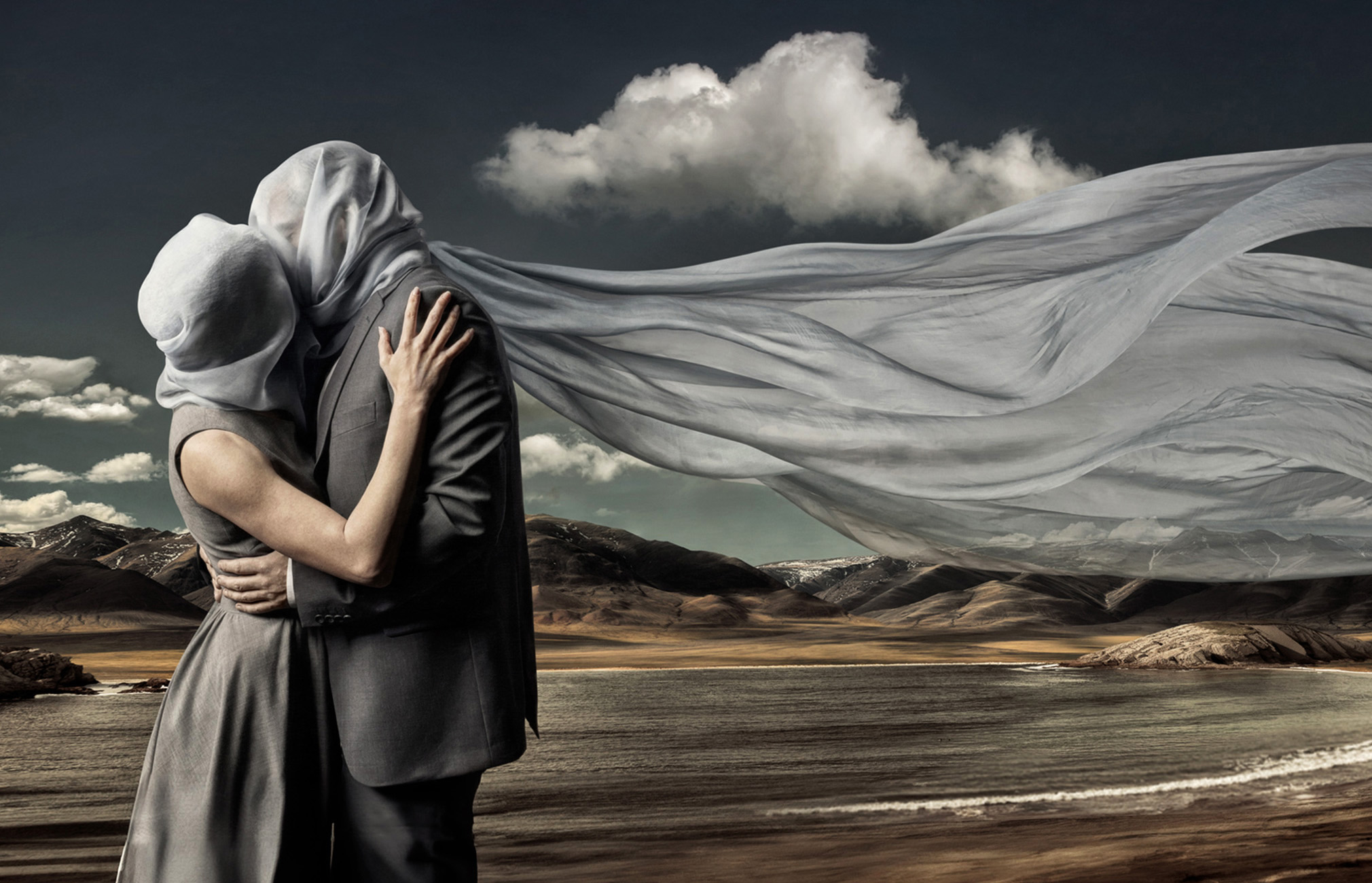 A couple embraces each other in a dramatic landscape, their faces are veiled, fabric blowing in the wind against a background of mountains and sky.