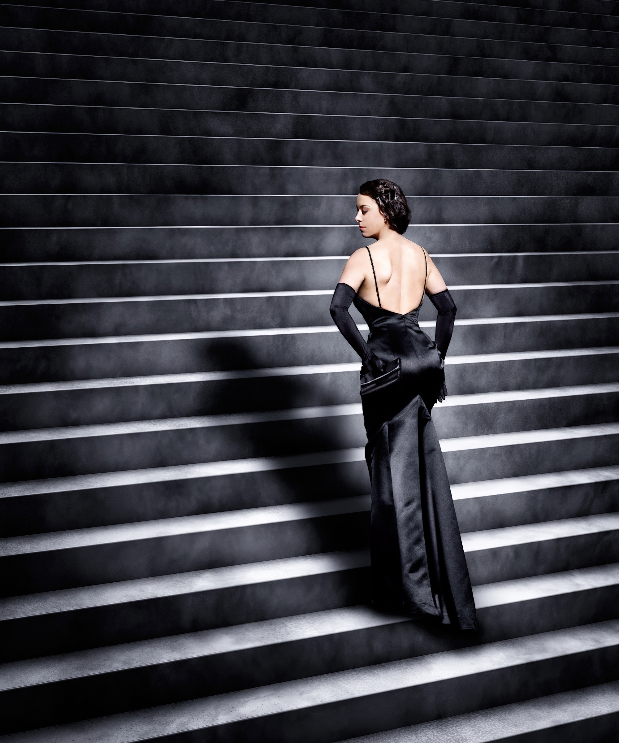 A woman in an elegant dress standing on a wide staircase with many steps. The individual is wearing an elegant, backless black dress. The staircase has sharp lines and shadows that create a pattern across the image. The photo is taken in black and white or has low color saturation, which gives it a dramatic and artistic expression.