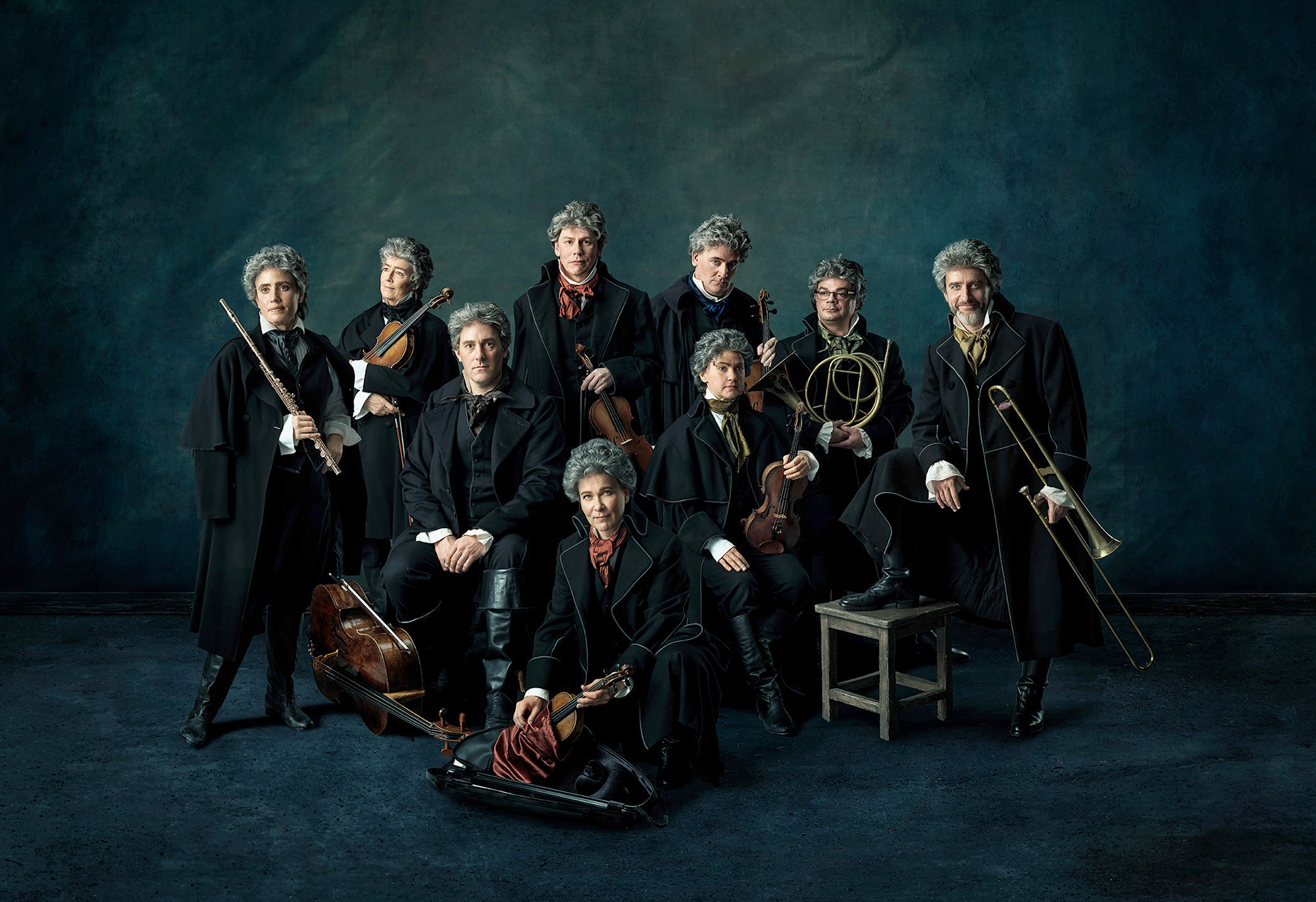A musical ensemble holding various musical instruments. The group consists of eight individuals posing against a dark, textured background. Each person is holding a musical instrument, including a violin, trumpet, and horn. They are dressed in formal black coats and trousers, with white shirts and neckties. One person is seated on a chair while the rest are standing or sitting on the floor. The lighting in the image is soft yet dramatic, giving the picture an artistic appearance.