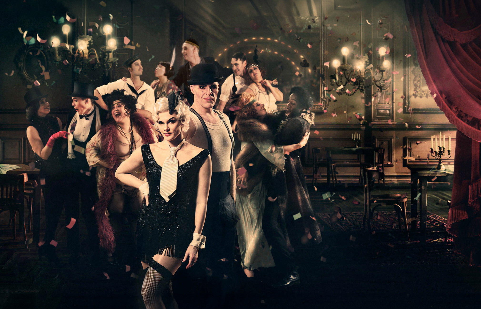 A group of people is in a festive environment. They are dressed in clothes that suggest an earlier era, possibly the 1920s. The background is a richly decorated indoor setting with dark wood tones and red draperies. Chandeliers and other light sources give the image a warm tone. The people seem to be engaged in dance or other festive activity, with flowers and other decorations visible in the background.