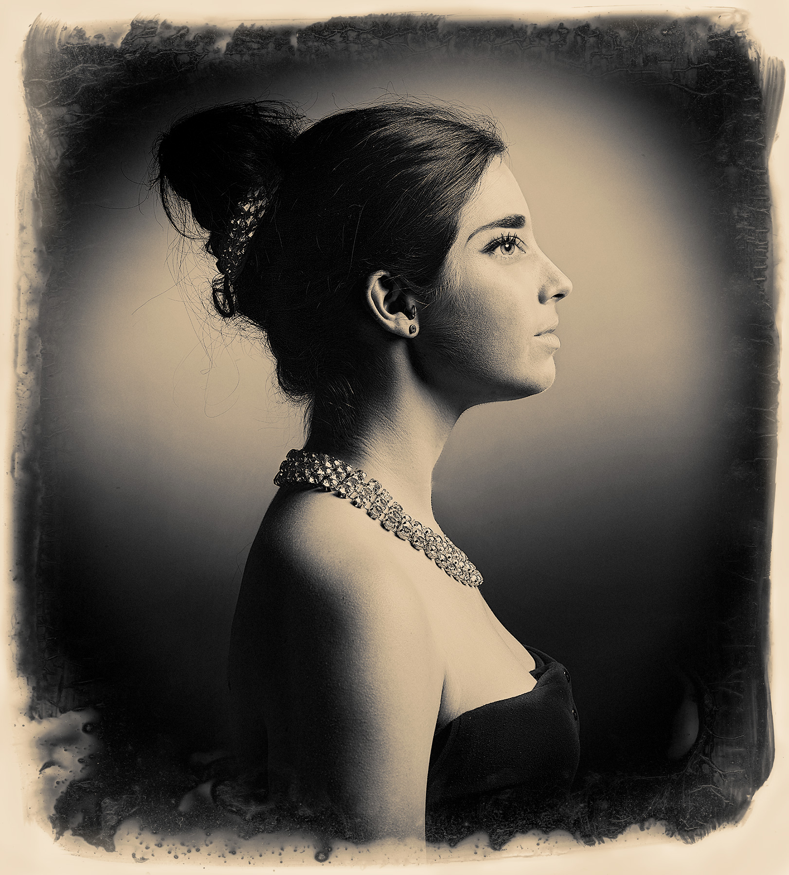 The woman in the picture is wearing a necklace and has her hair up. In black and white, the image offers a timeless and artistic expression. The background is blurred and nonspecific, drawing the viewer's focus to the person in the foreground. A detailed necklace worn around the person's neck adds a touch of elegance.