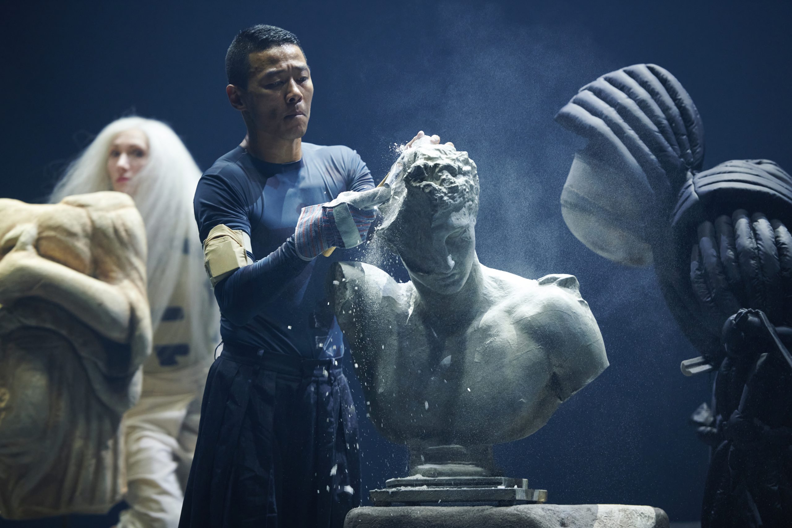 A man is carving a bust statue in a dark and dusty room. The person is wearing a long-sleeved blue shirt, gloves. The bust statue depicts a male figure with detailed facial features and musculature. In the background, more dancers can be seen, suggesting that this is taking place on a stage. The lighting in the room highlights the dust particles in the air and gives the scene an almost ethereal quality.