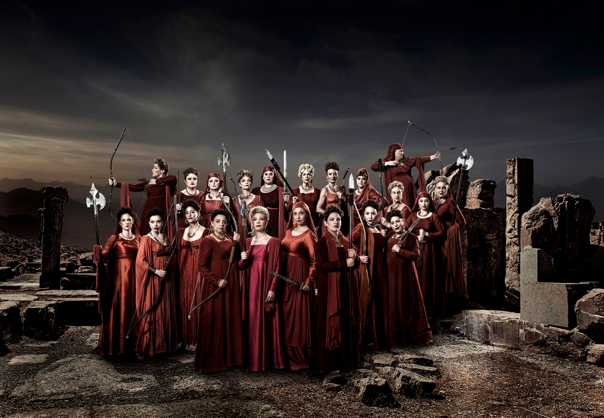 A choir donned in red robes, holding weapons; bows, swords and other medieval weapons are visible. They stand on rocky ground with ruins and a dark sky in the background. They are on an uneven, rocky surface that appears to be ruins. The backdrop presents a dark, stormy sky, creating a gloomy atmosphere.