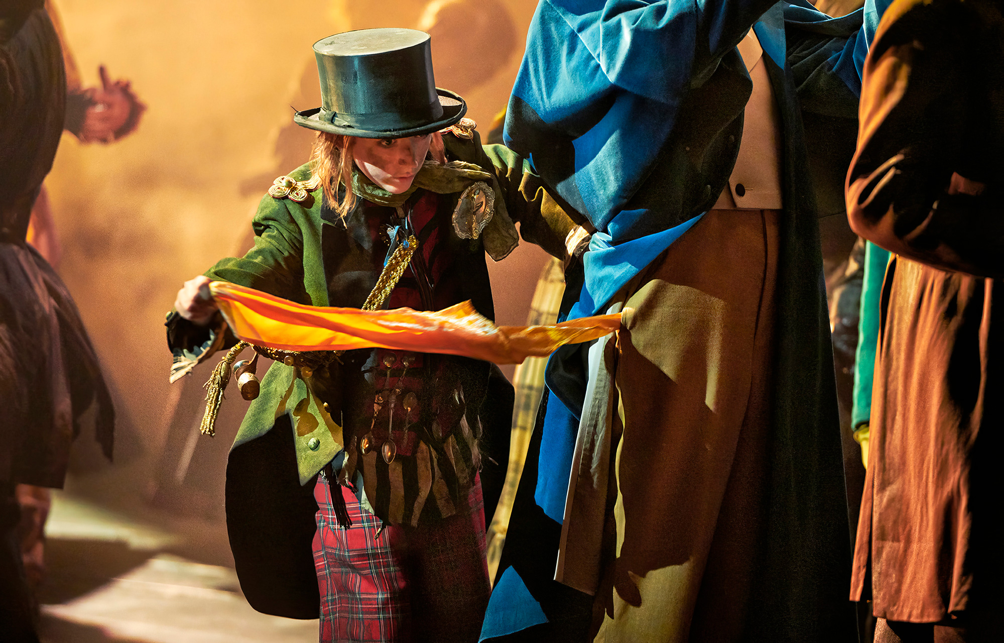 At the center stands a boy dressed in colorful clothing and wearing a hat. The individual holds a piece of fabric in orange and yellow. The person's face is hidden, but we can see that they are wearing a green jacket adorned with medals and red-checkered pants. A top hat adorns their head. Around the central figure, there are other people, also dressed in different costumes. Their attire includes blue and brown fabric. The background is misty, lending the picture a dramatic mood. The lighting is warm and casts shadows that highlight the details of the costumes and the environment.