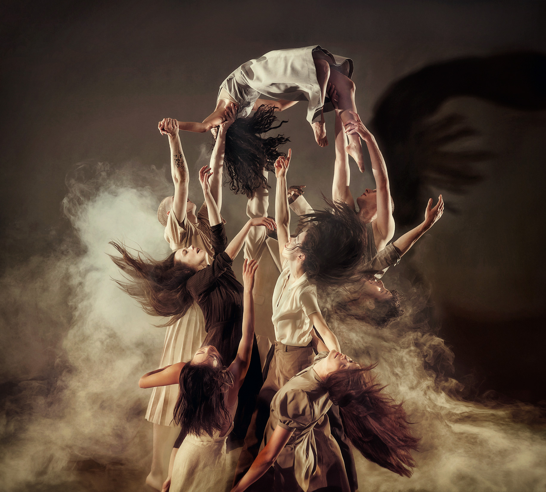 Dancers in a dramatic and artistic pose, surrounded by smoke. They are in various positions, some appear to be lifted or floating in the air. Their clothing is simple and in neutral colors, which contributes to the image's remote quality. The lighting is evocative and atmospheric, with shadows and highlights emphasizing the individuals' forms and the surrounding smoke. The overall mood in the picture is intense and emotional, conveying a sense of movement and energy.
