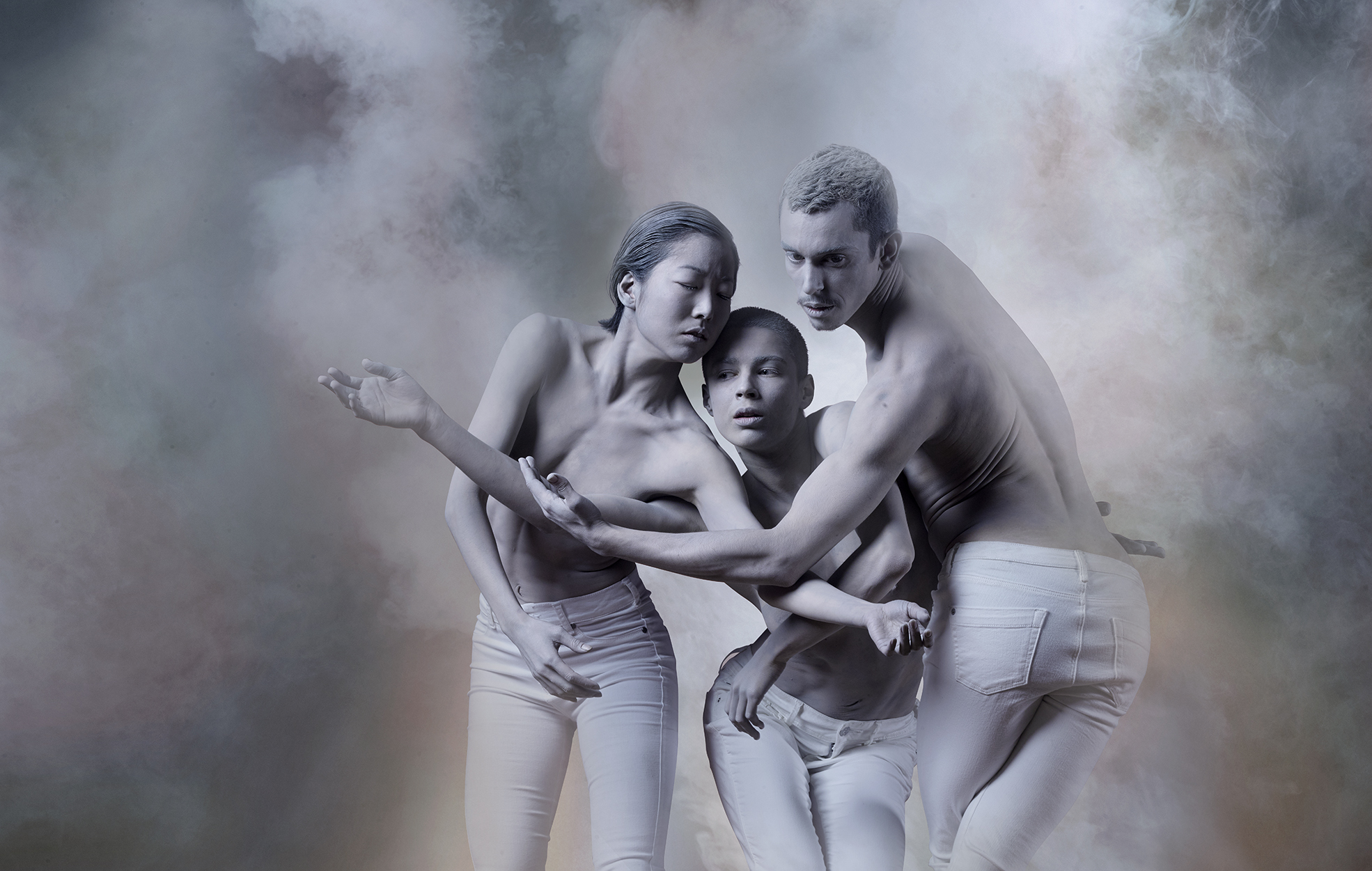 Three individuals with bare upper bodies engaged in a close embrace in a dance. They are wearing white trousers. The background is filled with a smoky or cloudy texture with various shades of gray.