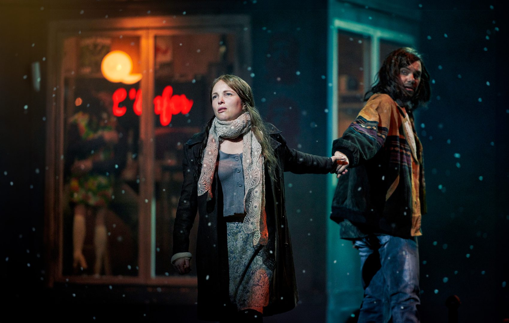 Two people walking outdoors at night, holding each other's hands. One person is wearing a scarf and a coat. The other person is dressed in a jacket with visible layers underneath, it's cold weather and snow is falling around them. In the background, a warmly lit window can be seen, displaying some decorations and the reflection of a red neon sign on a Café.