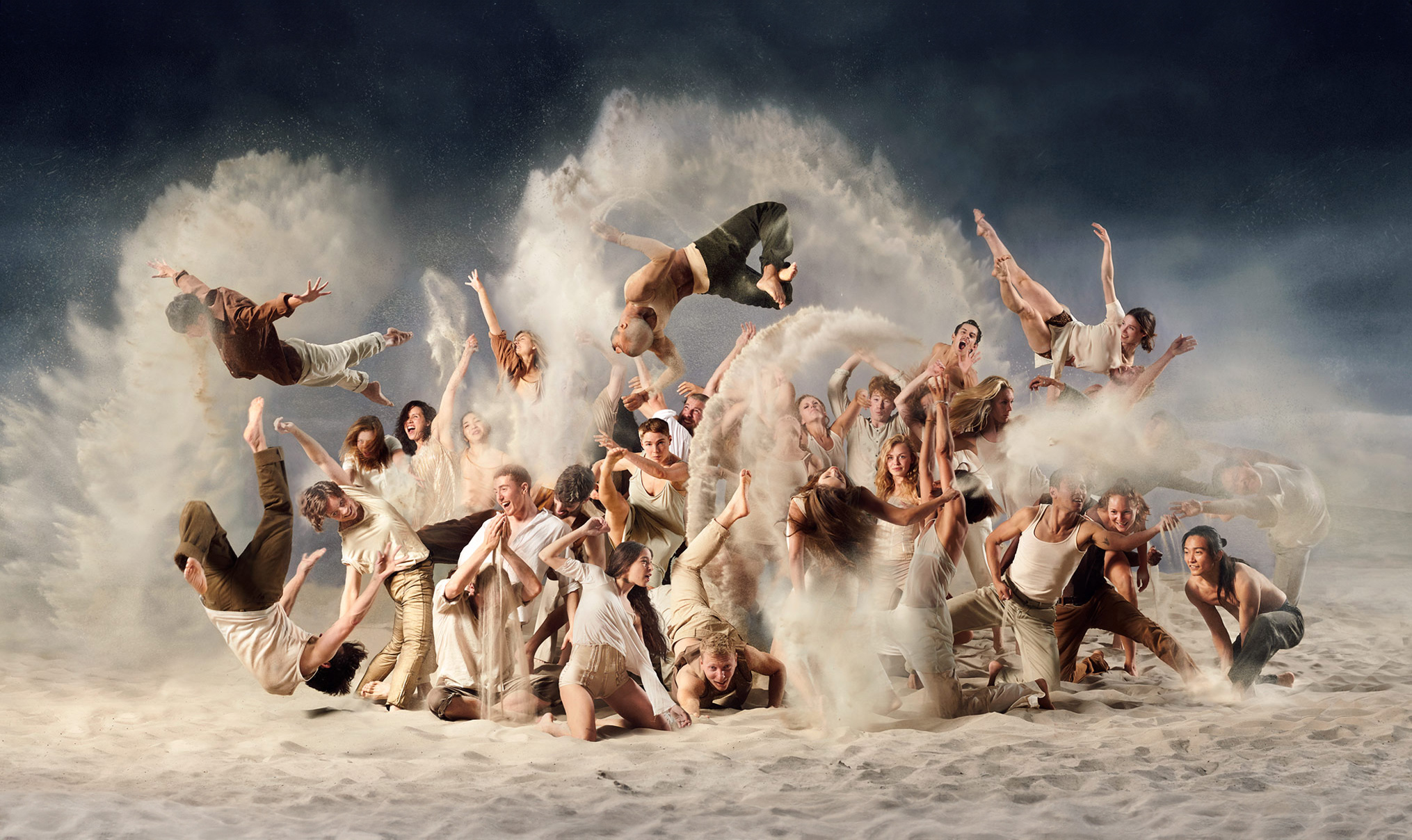 A group of people in various poses, surrounded by large clouds of sand against a dark background. The group is dressed in neutral colors, mostly beige and brown. Some individuals are jumping up, while others are bending or extending their arms. The thick cloud of sand creates a dramatic effect, and the background is dark, strongly contrasting with the bright clouds and the clothing of the individuals. There is no distinct environment; the focus is entirely on the people and the swirling sand.