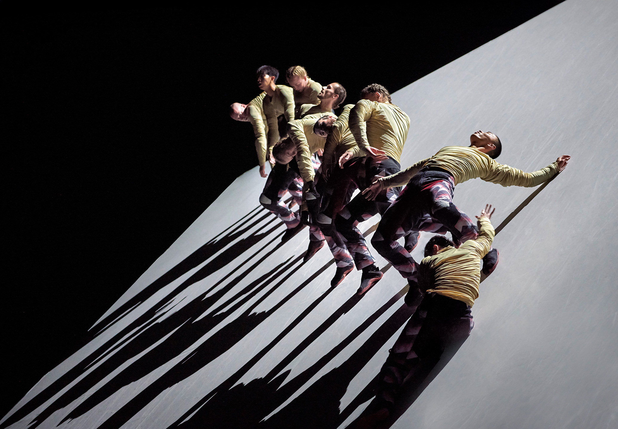 A group of dancers in yellow shirts and dark pants leaning against a sloping surface. They are positioned in various stances, creating an artistic and dynamic visual effect. Their elongated shadows are prominently emerging on the surface, adding a dramatic touch to the composition. The background is dark, which highlights the subjects and their shadows. The lighting is focused on the individuals, emphasizing their clothes and creating distinct shadows.