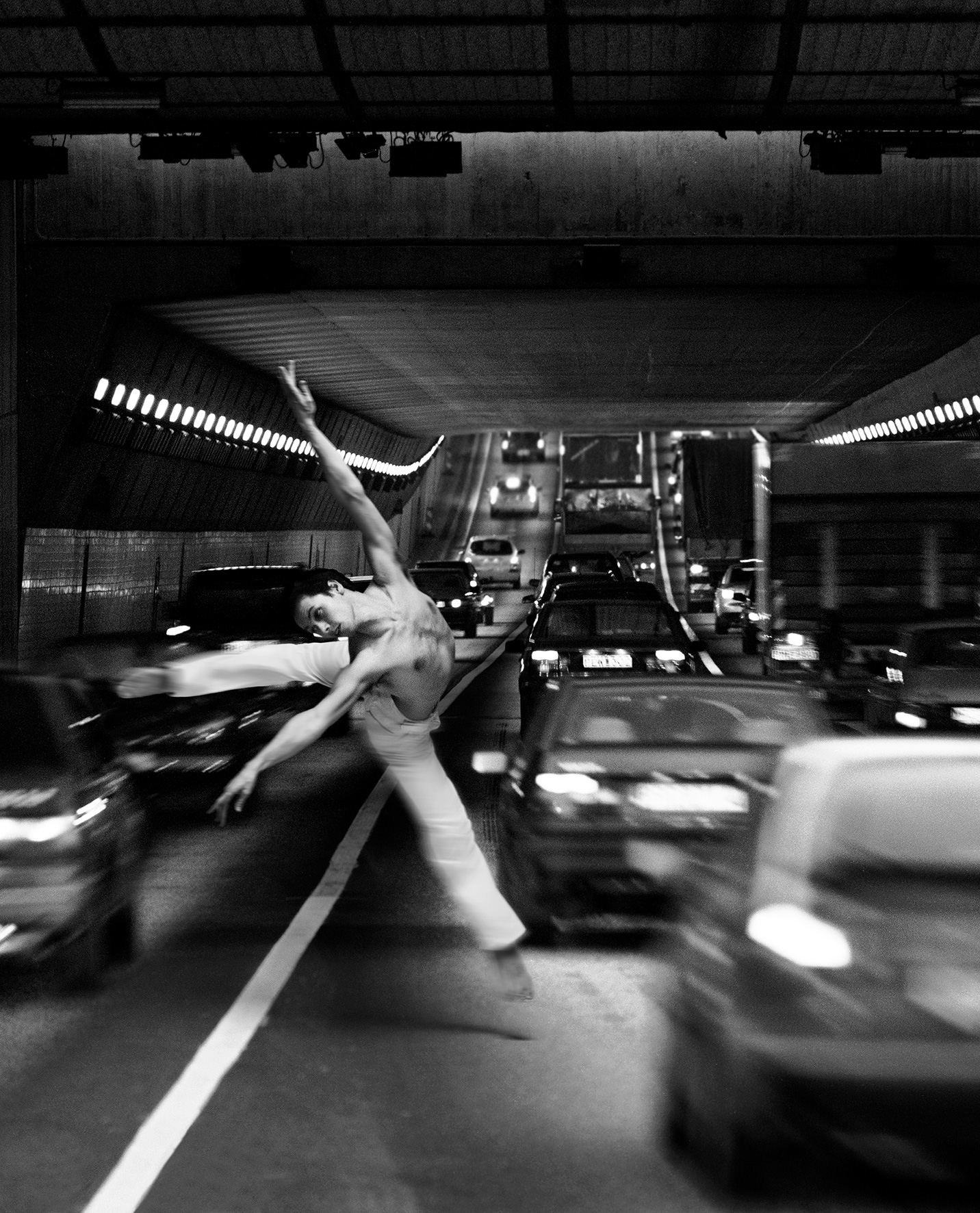 A person dancing in the middle of a busy road in the Tingstad Tunnel, Gothenburg. It's in black and white, capturing a dynamic and dramatic scene. The individual is in the air, seemingly dancing or jumping, and is positioned in the center of the road. Cars are moving around the person, creating a sense of motion and urgency. The tunnel is illuminated by ceiling lights, casting shadows and highlights that amplify the intensity of the moment. The cars are blurry, indicating motion.