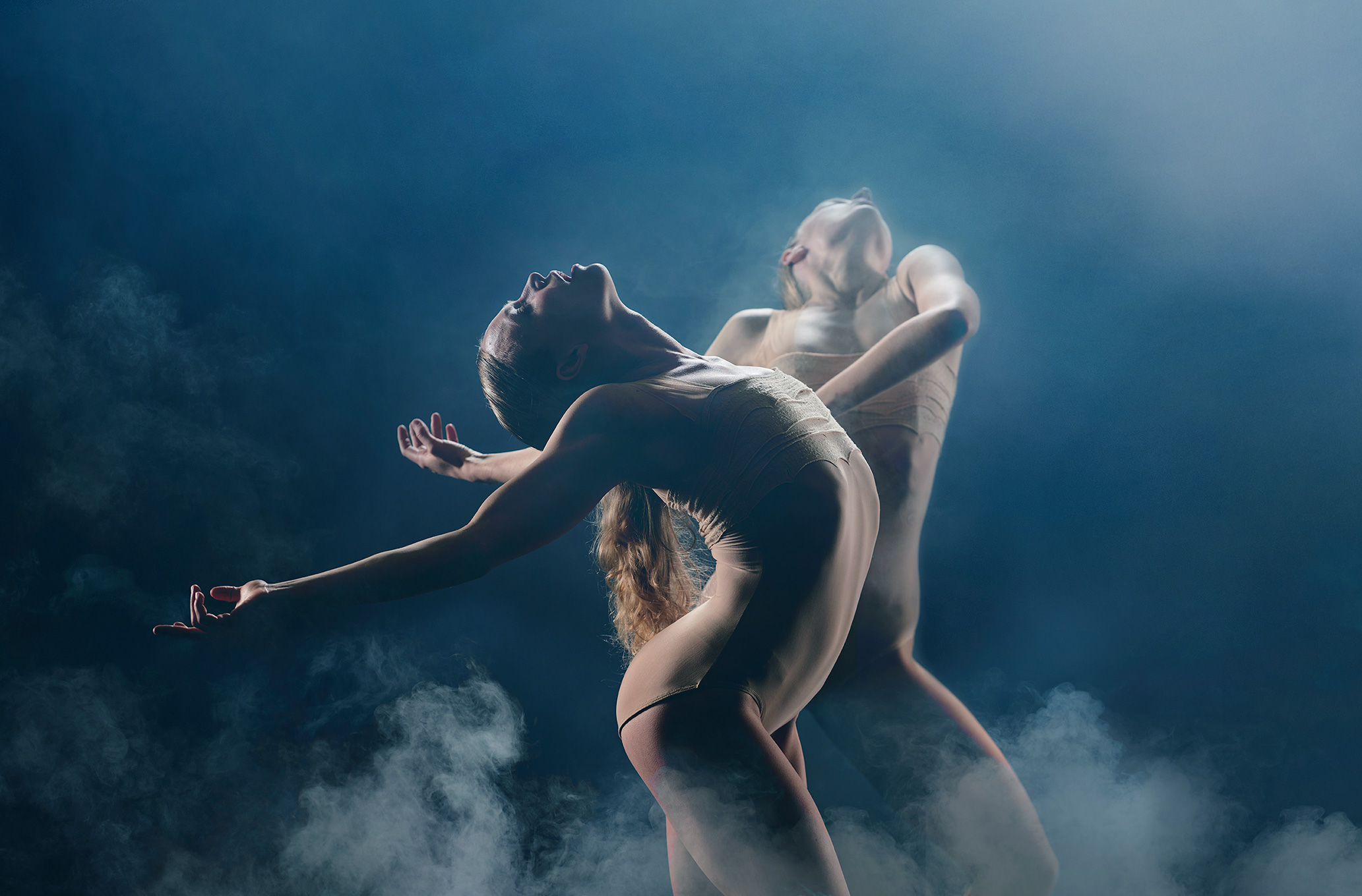 Two individuals in a dramatic dance pose surrounded by a cloud of smoke . They are illuminated from the side, casting parts of their bodies in shadow and highlighting their shapes against a dark background. The smoke lends a mysterious and surreal feel to the scene.