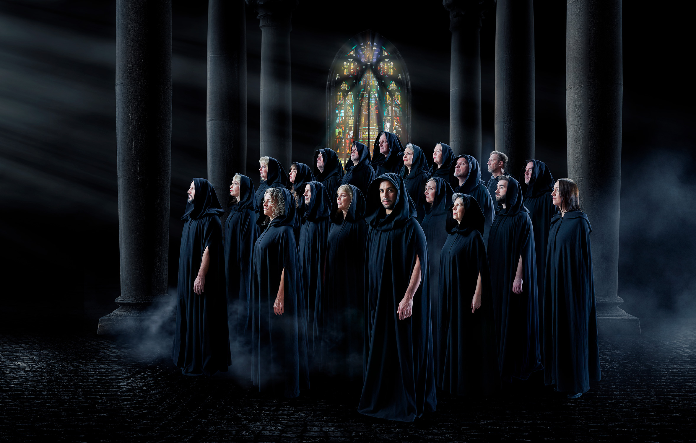 A choir dressed in black robes, standing in front of tall pillars and a colorful church window in the background. The atmosphere is dark and mysterious, and it appears they are located in a cathedral. Fog or smoke is visible at their feet, reinforcing the enigmatic mood in the image.