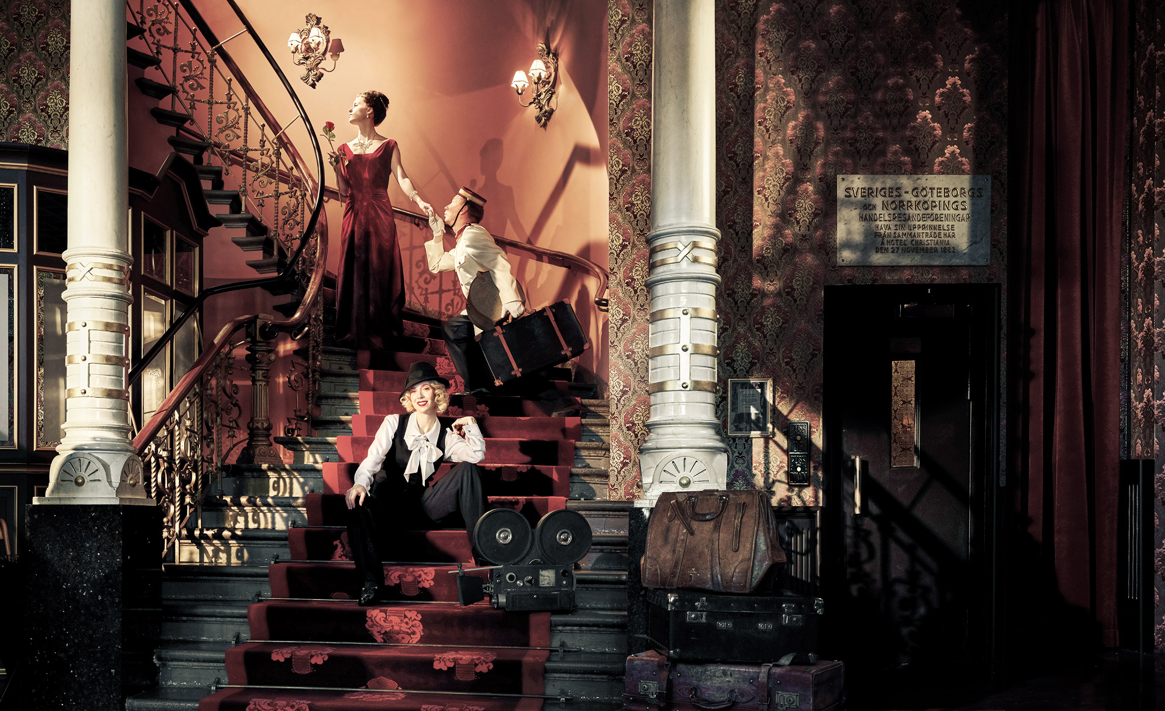 An indoor scene from the early 1900s featuring two individuals dressed in period-appropriate clothing. One person is seated at the foot of an elegant staircase, clutching an umbrella, while the other stands higher up on the stairs with luggage near them. Vintage suitcases are scattered around the area, and the walls are festooned with floral patterns and artwork. The scene gives the impression of a hotel reception with rich history.