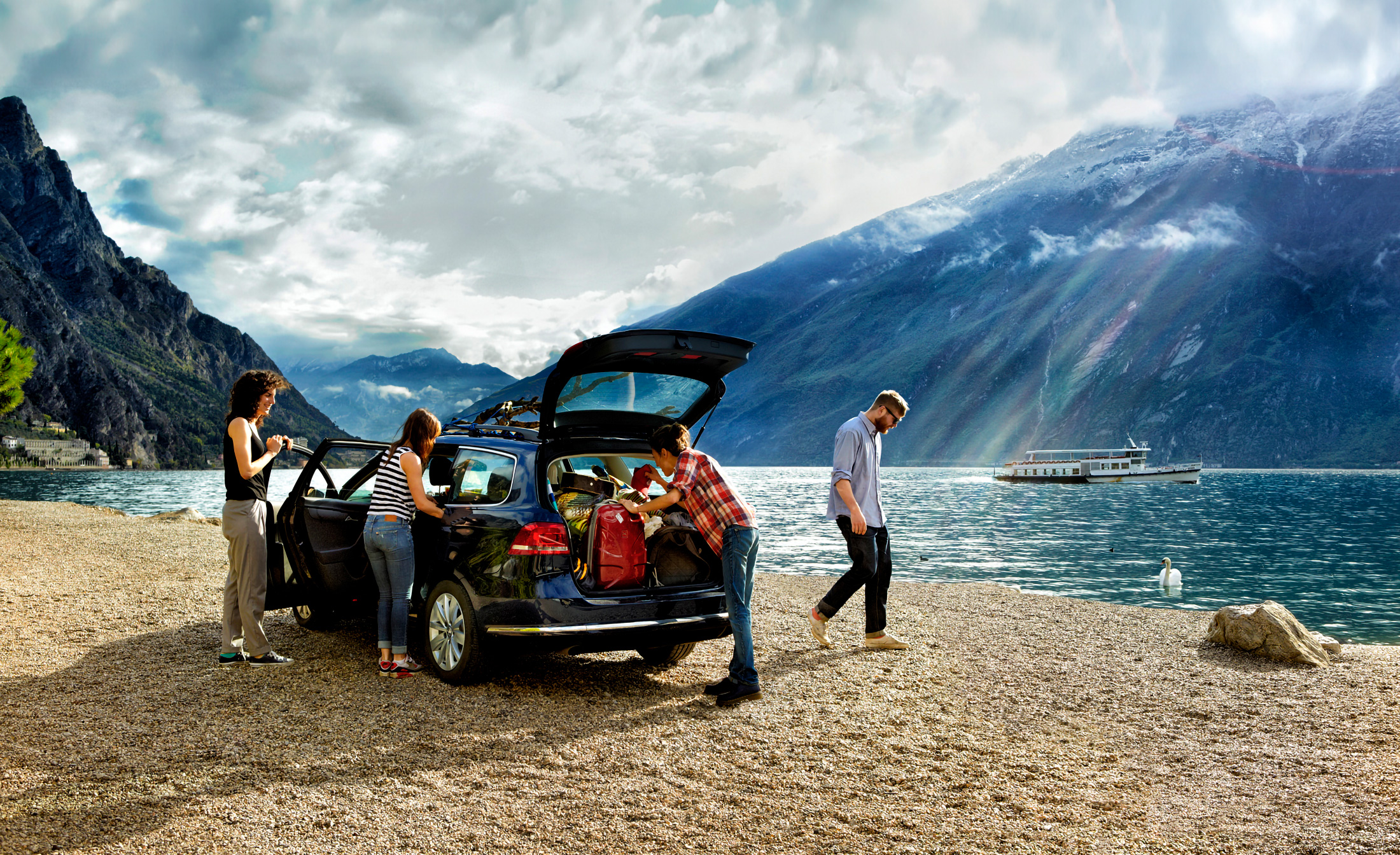A scene by a lake with mountains in the background. There is a car parked on a gravel beach, and several people seem to be busy unloading the car. A man is walking towards the water, and two women are standing next to the car's open trunk. It's sunny weather with a few clouds, which gives a soft light over the scene, a ship can be seen in the distance on the water. It appears to be a relaxing day outdoors with friends.