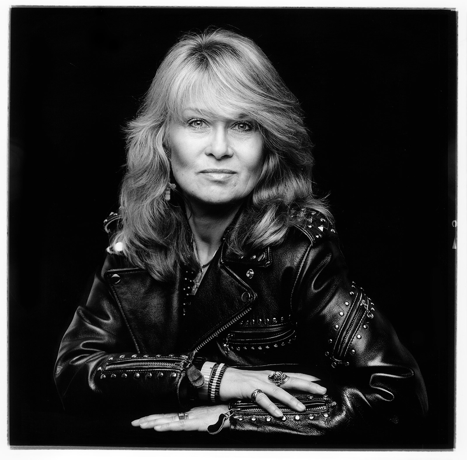 A black and white portrait of a woman. She wears a studded leather jacket, which gives off a rock-and-roll style. Her hair is long and in light in color, cascading over her shoulders. Her hands rest on a table, adorned with bracelets and rings, enhancing the style of her outfit. The background is completely black, accentuating the subject.