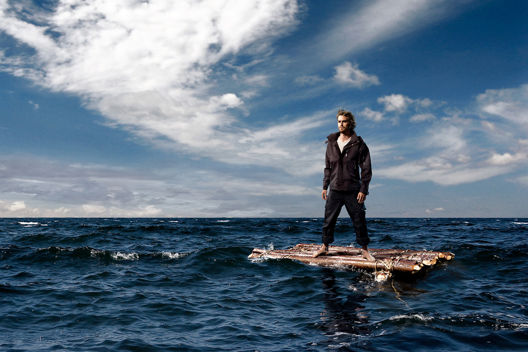 A person standing on a homemade raft in the middle of a body of water, which appears to be the sea. The raft is made of what looks like wooden planks and barrels. The person is wearing dark trousers, a jacket, and shoes. Above, the sky is partly cloudy with sunlight streaming through the clouds, creating a dramatic effect.